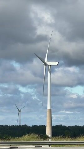 The video shows a close-up view of a wind turbine with a concrete base. In the background, another wind turbine is visible, along with a line of trees and a cloudy sky. A black SUV drives past the wind turbine.