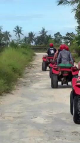The video shows a group of five people riding red ATVs down a dirt path. The camera follows the group from behind. All riders are wearing red helmets. The rider in the front is wearing a red t-shirt and the rider in the back is wearing a black jersey with the number 16 on the back. The path is bordered by green vegetation and palm trees. The sky is cloudy.