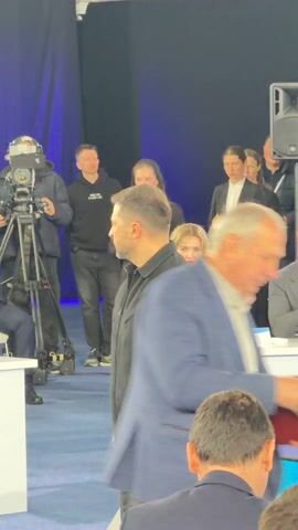 The video captures an indoor event, likely a conference or formal meeting, with a dark blue backdrop. In the foreground, the backs of two men in dark suits are visible, seated at a table with a glass of water and a bottle. A partially visible white table tent shows the letters "ГРИ" and "Д."

In the mid-ground, a man in a black shirt and pants stands facing away from the camera. A woman in a black blazer, black pants, and white sneakers walks from right to left, holding a red folder or book. She has a slight smile. Another woman with blonde hair is seated to the right of the standing man.

Further in the background, a camera on a tripod is filming, operated by a person behind it. Several other individuals are visible, mostly dressed in business attire, some standing and some seated. The overall impression is an organized event with participants moving between tables or preparing for a session.