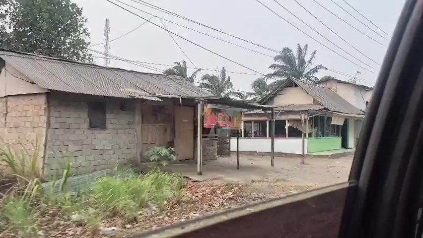 The video shows a view from inside a vehicle looking out at a street scene. On the left side of the screen, there is a small building with a colorful advertisement on its side. In the center, there are several small shops or dwellings with simple architecture and corrugated metal roofs. Some have awnings or covered areas in front. There are palm trees visible in the background. Several motorcycles are parked along the street. Some people are visible outside one of the buildings, sitting at tables. The ground is unpaved and dusty. The overall impression is of a modest, perhaps rural or semi-urban, commercial or residential area. The sky is overcast.