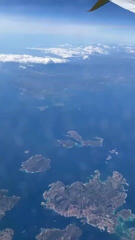 An aerial view from an airplane window shows a vast expanse of deep blue sea dotted with numerous islands and islets, characteristic of the La Maddalena archipelago near Baja Sardinia, Italy. The rugged coastlines of the islands are visible, some featuring patches of urban development. In the far distance, a mainland coastline with mountains can be seen under a clear blue sky, with a line of white clouds resting on the horizon. The tip of a yellow airplane wing is visible in the upper right corner of the frame, indicating the vantage point. The scene depicts a sunny day, showcasing the beautiful natural landscape from above. No people or visible text are present in the video.