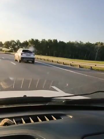A white SUV is traveling on a highway in the right lane. The camera is positioned inside another vehicle, looking out the windshield. The SUV is in front of the camera vehicle and appears to be driving in the same direction. The sun is low in the sky, casting a warm light on the scene. Trees line the side of the highway.