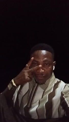 A man is centered in the shot against a black background. He wears a striped, zip-up shirt with a tan collar, a gold watch on his left wrist, a ring on his left hand, and white wireless earbuds. He makes a peace sign with his left hand initially, then sticks out his tongue briefly before smiling slightly. The lighting is dim, illuminating the man's face and clothing. The background appears to be completely dark.