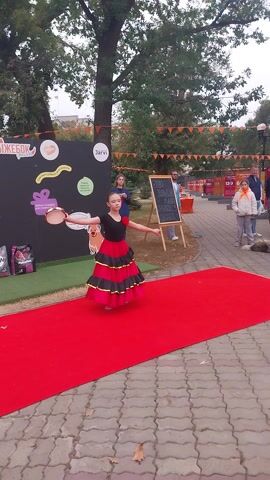 The video shows a young girl performing a dance on a red carpet in Kargala, Russia. She is wearing a black leotard with a red and black ruffled skirt and white socks. She is holding a tambourine and appears to be dancing to music.

In the background, there is a black board with some text written on it, which is difficult to read. There are also several posters and decorations on a black backdrop behind the girl. One poster has the text "Jarvi" on it. Further in the background, there are other people watching, including adults and children. Some are standing and some are sitting. The overall scene appears to be an outdoor event, possibly a festival or a performance. The weather seems overcast, suggesting it might be late afternoon or early morning. The ground is paved with interlocking concrete blocks. Trees are visible on the left side of the frame. The overall mood seems festive, with the performer focused and engaged in her dance.