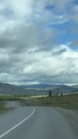 The video begins with a view from a vehicle driving down a paved road. The road curves to the left, with a white solid line marking the edge. Rolling hills and mountains form the backdrop, with a cloudy sky overhead. As the vehicle continues, a red truck comes into view, driving in the opposite direction. The truck is a large, modern semi-truck with a white cab and a red trailer. The truck passes the vehicle, and the road continues onward through a valley. There are power lines visible on the right side of the road, and various signs are posted. The landscape appears to be rural and somewhat undeveloped. The video is shot from a first-person perspective, as if the viewer is a passenger in the vehicle.