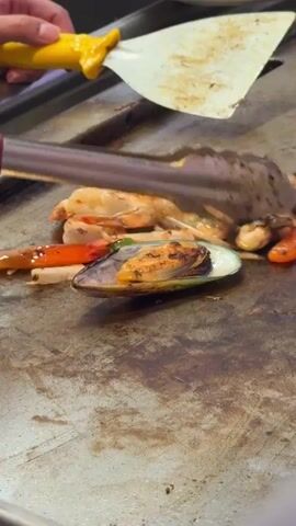 A close-up shot shows a grill where seafood and vegetables are being cooked. In the foreground, a large mussel is visible. To the left, shrimp and other seafood are being tossed with tongs. A spatula with a yellow handle is also being used to move the food. Steam rises from the hot grill. In the background, a white plate holds raw red meat. The grill surface is dark and appears well-used. A hand with a yellow utensil is on the upper left side of the frame.