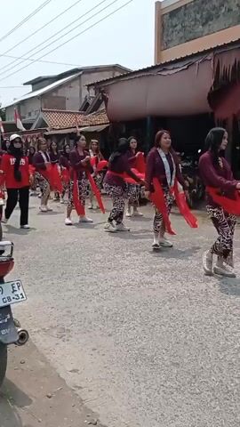 A group of women in maroon tops and patterned skirts with red sashes are marching in a parade. Some are holding red scarves. One woman in the front is on a red scooter with a child. Another woman in a red sweatshirt and black pants walks ahead of the main group. There are buildings and trees in the background, and the ground is paved. Another scooter is partially visible on the left. The overall scene appears to be a street celebration.