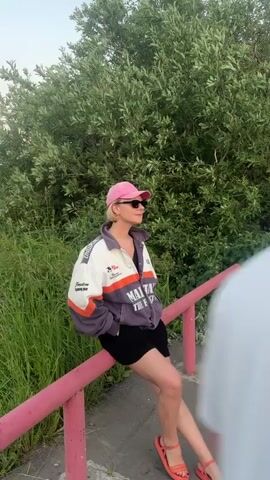 A person wearing a pink baseball cap, sunglasses, a purple and white jacket, and black shorts sits on a pink railing next to a body of water. The person is looking to their right, with their legs crossed. The water is calm and reflects the surrounding trees and sky. The sky is a pale blue with hints of pink and orange. The scene is set in a natural environment, with lush green vegetation. The video pans across the water, showing a wider view of the landscape.