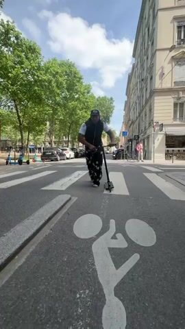 A man performs a scooter trick on a sunny day in Lyon, France. He rides a black kick scooter along the edge of a raised curb, then executes a jump off the curb, landing back on the asphalt road. He is wearing a black vest over a white t-shirt, black and white patterned baggy pants, and a baseball cap.

The setting is an urban street with tall green trees lining a promenade on the left. Buildings with classical architecture are visible on the right side of the street. The road features a painted white bicycle symbol, indicating a bike lane. In the background, there are parked cars, several light blue shared electric scooters, and other pedestrians. The sky is bright with scattered clouds. Street signs, including a blue circular sign with a white arrow and a blue rectangular sign with a white bicycle symbol, are also visible.