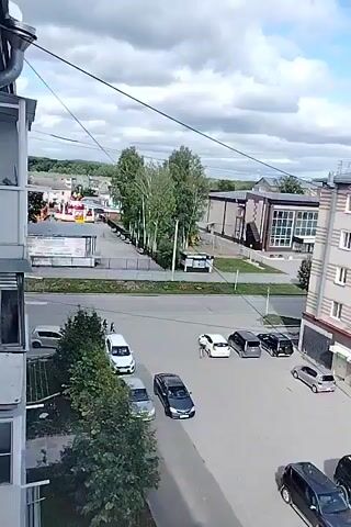A drone shot moves over a residential area with multiple buildings and trees. Several cars are parked in a lot, and a person is walking on a sidewalk. The sky is partly cloudy.