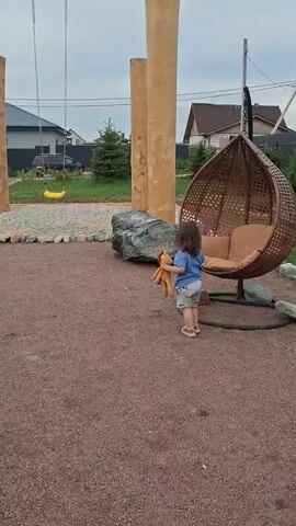 A young child walks across a gravelly playground holding a stuffed lion. The child is wearing a blue t-shirt and shorts and pink shoes. Behind the child, there is a large, hanging chair and a swing set made of wooden posts. In the background, there are houses and trees under a cloudy sky. The child stops and looks at the camera, then continues to walk.