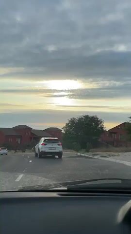 This media file captures a car's-eye view of a road trip in Aktau, Kazakhstan. The primary subjects are the road, several cars, and modern houses with red roofs situated on a hillside. The scene is set at dusk, with the sun breaking through a cloudy sky, casting a warm glow. The weather appears to be mild, with the clouds suggesting a potentially cooler evening.

The activity depicted is driving along a paved road that curves and descends towards a body of water, likely the Caspian Sea, visible in the distance. The road has clear white lane markings. Two white SUVs are prominently featured, one ahead of the viewer's car and another further down the road. Several other vehicles are visible in the distance, indicating ongoing traffic.

Notable details include the architecture of the houses, which appear to be contemporary and somewhat luxurious. The vegetation around the houses is sparse, consistent with a coastal desert environment. The sky displays dramatic cloud formations and a vibrant sunset, creating a serene and picturesque atmosphere. The time of day is clearly indicated by the low angle of the sun and the soft light. There is a license plate visible on the white SUV in the foreground, which appears to read "C310YK 157". The overall emotion conveyed is one of a peaceful journey through a scenic landscape as day transitions into night.