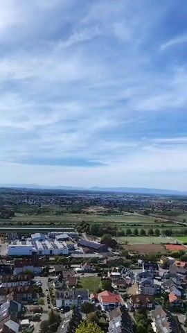 The video pans across a landscape under a blue sky with scattered clouds. In the foreground, a few trees with green and brown leaves are visible. Below the trees, a residential area with numerous houses, many featuring red-tiled roofs, stretches out. Roads and small yards are interspersed among the houses. Further in the distance, there are some industrial buildings with white and grey roofs. Beyond the buildings, a wide expanse of green fields is visible, with a few trees scattered throughout. A winding river or canal is also present in the middle ground. In the far distance, a hazy, mountainous horizon can be seen, with a town or city spread across the plain. The overall impression is of a rural or suburban setting with a vast, open landscape in the background.