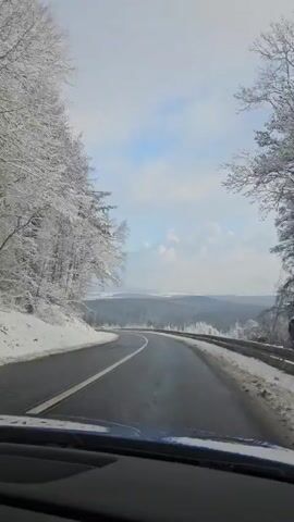 A car travels on a black asphalt road through a snowy winter landscape. The trees lining the road are covered in snow. The sky is light blue with some white clouds. Another car appears in the distance, coming from the opposite direction. The road curves to the left. A guardrail is visible on the right side of the road.
