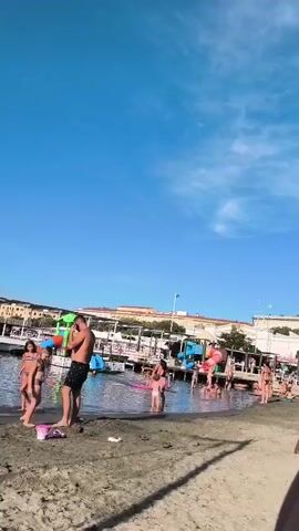 This is a landscape video filmed at a beach on a sunny day. The sky is a clear, bright blue with a few wispy white clouds. The foreground is a sandy beach with some debris scattered on it. In the midground, a shallow body of water meets the sand. Several people are visible, mostly children and a few adults, in swimwear. Some children are playing in the shallow water, while others are on the sand. One man stands in the water, appearing to talk on his phone. A pink bucket lies on the sand near him.

Further back, a pier or boardwalk extends into the water, lined with various inflatable structures and possibly some beach amenities or stalls. Several people are gathered around these structures. In the background, a line of buildings with terracotta roofs suggests a coastal town. The overall atmosphere is one of a relaxed summer day at the beach, with people enjoying the water and sunshine. The time of day appears to be midday or early afternoon, given the bright sunlight casting shadows. There is no visible text in the video.