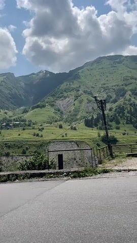 The video shows a winding asphalt road through a lush green mountain valley. The road curves to the left, with trees on the left side of the frame and a steep, terraced mountainside on the right. Small buildings are visible scattered across the hillside in the distance. The sky is blue with scattered white clouds, and the sun appears to be shining. There is a utility pole on the right side of the road.