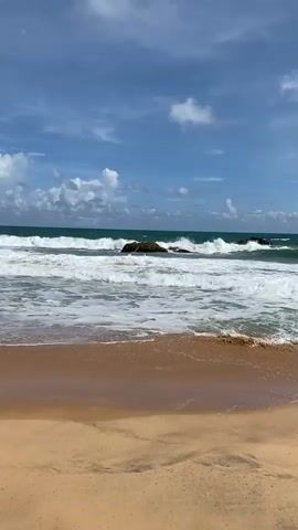 A wide shot captures a serene beach scene with a dynamic ocean. The foreground features wet sand, indicating the tide has recently receded. A gentle curve of sand extends across the bottom of the frame, with subtle ripple marks suggesting the movement of water. In the middle ground, the ocean is a vibrant turquoise, with white foamy waves rolling towards the shore. Two large dark rocks are visible in the water, with waves crashing around them, creating a dramatic contrast between the static rocks and the fluid motion of the sea. A few smaller rocks are also scattered throughout the water. The horizon line is clear, separating the ocean from a vast blue sky. The sky is dotted with fluffy white clouds, some of which are illuminated by sunlight. The overall impression is one of natural beauty and tranquility.