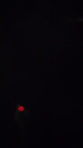 The video begins with a dark sky and a distant light, possibly a streetlamp. A small, blurry, light-colored object appears in the upper right quadrant. It is then obscured. A faint streak of orange light, possibly a firework, is seen briefly. More fireworks begin to explode in the sky, with various bright bursts of light and sparks against the blackness. Some lights appear green, others orange, and some white. Another blurry object, possibly smoke or debris, is visible in the upper right. The fireworks continue to explode in different patterns and intensities. The video ends with a few small, distant lights appearing and disappearing.