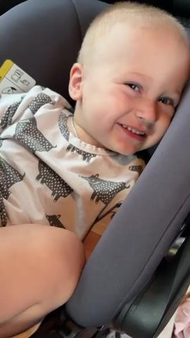 A toddler is visible in a car seat. The toddler is wearing a white t-shirt with a black fox print. The toddler is smiling and looking at the camera. The car seat is gray and is in a car. There is a hand visible in the top left corner of the frame, briefly touching the toddler's head. The video is shot from a close-up perspective.