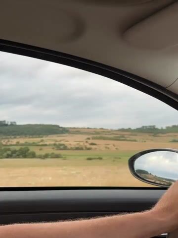 The video shows a view from inside a car traveling through a rural landscape. The camera is positioned to look out the side window. The foreground shows a person's arm and hand resting on the car door. In the middle ground, a field of dry, golden grass stretches across the landscape, with a side mirror visible reflecting the sky and some of the scenery. The background consists of rolling hills covered in greenery and patches of cultivated land. The sky is overcast with dramatic clouds. The overall impression is of a journey through the countryside on a cloudy day.