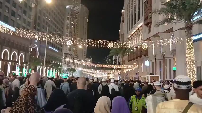 The video shows a large crowd of people walking down a street at night. The street is decorated with strings of lights, some shaped like stars and crescents. Tall buildings line both sides of the street, some of which are lit up with white and green lights. The people in the crowd are diverse, with some wearing traditional Muslim clothing, while others are wearing more Western attire. Some people are carrying bags or backpacks. The overall atmosphere is bustling and festive.