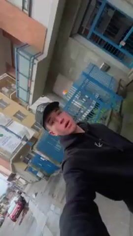 The video shows a young man taking a selfie video while walking down a wet sidewalk in an urban area. He is wearing a black hoodie and a baseball cap.  The camera moves as he walks, showing buildings, cars parked along the street, and a row of garbage and recycling bins of different colors. The sky is overcast.