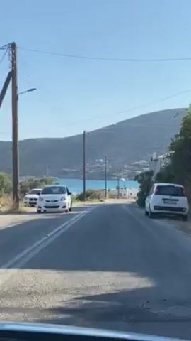 The camera faces forward and rolls down a paved, gray road towards a blue bay with several sailboats anchored near the shore. On either side of the road are parked cars and white-washed buildings typical of a Greek island setting. Power lines run parallel to the road, supported by wooden poles. A grassy hillside frames the view behind the bay. The sky is clear and blue.