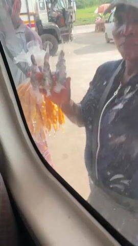 The video shows a view from inside a vehicle, looking out the side window. Two vendors approach the vehicle. The vendor to the left is holding a large plastic bag filled with orange-colored snacks, possibly fried plantains or similar food items. The vendor is wearing a long-sleeved shirt, patterned pants, and a head covering. The vendor on the right is also close to the window, visible from the waist up. They are wearing a dark shirt with light-colored stains and a strap across their chest, possibly from a bag. In the background, there is a large white truck with a driver visible in the cab. Other vehicles are parked nearby, and there is a person under an umbrella in the distance. The ground appears to be concrete or paved.
