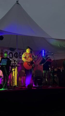 The video shows a live music performance on an outdoor stage under a large white tent. Three musicians are visible. On the left, a woman wearing a red and white headband plays a keyboard. In the center, a man in a hat and a colorful shirt plays an acoustic guitar and sings into a microphone. On the right, a person wearing a cowboy hat plays the violin. The stage is lit with colorful LED lights, including purple, blue, and green. A sign behind the musicians reads "LOFE ARTKO". The sky in the background is dark, suggesting it is evening or nighttime.