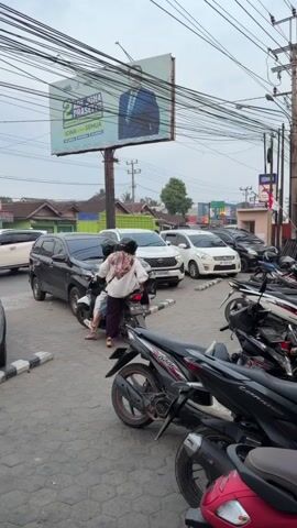 A street scene shows several parked motorcycles and cars. A billboard featuring a man in a suit is visible overhead. Two people are on a motorcycle. One person is driving, and the other is sitting behind them, facing away from the camera. The person in the back is wearing a white jacket with a colorful design on the back and dark pants. The driver is wearing a dark shirt and helmet. The motorcycle is stopped, and the rider in the back appears to be looking towards the parked cars. The environment suggests a commercial or public area, with buildings and power lines visible.