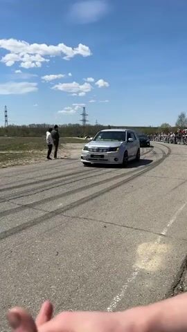 The scene is an outdoor gathering on a paved road, on a bright, sunny day with scattered clouds. There are multiple cars performing donuts, leaving tire tracks on the asphalt. A small crowd of people is gathered on the right side of the road, observing the activity. In the foreground, a person's hand is partially visible. A light grey car with modifications is pulling away from the camera, and a black car is actively drifting and creating tire marks. A man in a grey jacket is walking along the edge of the road, away from the crowd. In the background are power lines and a distant treeline.