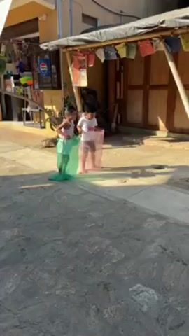 The video depicts two children participating in a sack race. The child on the left has a green sack, and the child on the right has a pink sack. They are racing on a paved area that transitions to cobblestone. In the background, there is a building with a small store and another structure with wooden walls and a makeshift roof adorned with colorful flags or cloths. The scene is illuminated by bright sunlight.