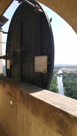 The video shows a view from a high vantage point, looking out through an arched opening. A large, dark, circular object, possibly a dish antenna or a bell, dominates the foreground and partially obscures the view. It is mounted on some kind of mechanism, with wires and brackets visible. Below the circular object is a stone ledge or parapet, with an arrow marking visible on its surface. Beyond the opening, a river or canal can be seen, lined with trees, and in the distance, there are fields and a hazy horizon. The sky is clear and blue. The lighting suggests it is daytime.