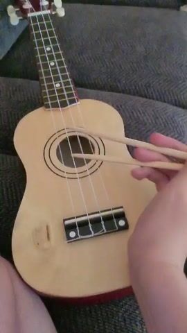 A person's hands are visible in the foreground, holding two wooden drumsticks. The drumsticks are being used to strike the strings of a light-colored ukulele, which rests on a dark gray, textured surface, possibly a couch. The ukulele has four white strings and a circular soundhole with concentric black rings. The neck of the ukulele, with frets and tuning pegs, is visible extending upwards. The action appears to be rhythmic striking of the ukulele strings with the drumsticks.
