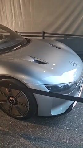 The video pans across a silver Mercedes-Benz EQXX car parked indoors. The car is behind a velvet rope barrier, with stanchions visible in the foreground. The lighting suggests an indoor exhibition or showroom. The car's sleek design, distinctive wheels, and the "EQXX" badging are prominently featured. There are no people visible in the frame. The background appears to be a white curtain or wall.