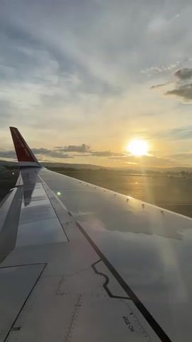 The video shows a view from an airplane window during what appears to be either takeoff or landing. The sun is setting or rising, casting a warm glow across the sky and reflecting off the airplane's wing. The wing is in the foreground, and its details are visible, including rivets and markings. The landscape outside the window is blurred due to motion, but suggests a runway and some distant hills or mountains. The sky is partly cloudy, with streaks of cirrus clouds visible. The overall impression is one of flight and a beautiful natural spectacle.