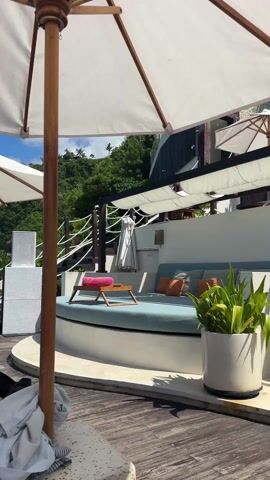 The video shows a bright, sunny outdoor lounge area with comfortable seating under white umbrellas. The seating consists of blue cushions arranged on a white base, accessorized with orange and grey pillows. Two small wooden tray tables hold rolled pink towels. A large potted plant stands next to one of the seating areas. Wooden decking covers the ground. In the background, there is lush greenery. Gentle music plays.