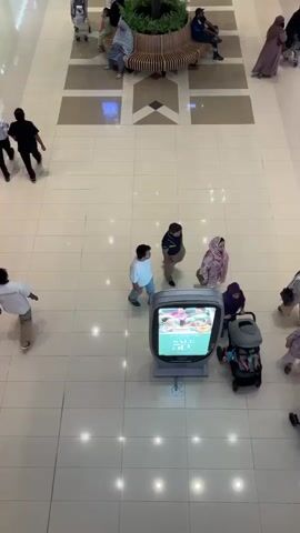 The video shows an overhead view of a busy indoor shopping mall. In the foreground, a digital display screen shows an advertisement for a sale. Several people are visible walking around the mall, including a man pushing a stroller with a child, a couple of children with an adult, and a lone man walking away from the camera. In the background, several people are sitting on a curved bench around a potted plant, and others are walking through the mall. The mall floor is tiled, and there is a decorative patterned walkway in the center of the frame.