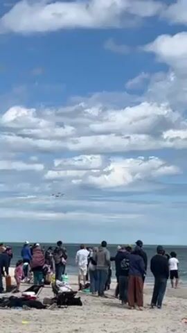 Here is a concise description of the video:

The video captures a scene at a beach, with a crowd of people watching an air show. Four airplanes are visible in the sky, flying in formation and leaving smoke trails behind them. The sky is blue with puffy white clouds. The beach is sandy, and there are gentle waves rolling in. The crowd consists of men, women and children, all looking up at the planes.