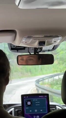 The video captures a view from inside a car, looking forward through the windshield and at the rearview mirror. A person's head and shoulder are visible in the foreground on the left, suggesting they are either driving or a passenger. The rearview mirror reflects the face of another person, with their eyes clearly visible and looking forward. Through the windshield, a winding road is visible, bordered by lush green vegetation, indicating a rural or mountainous setting. The sky appears overcast, suggesting a cloudy day. The car's dashboard features a modern infotainment screen displaying music playback information. The text on the screen indicates "Music," "Album," and "Artist," along with playback controls. The overall scene suggests a journey through a scenic, possibly mountainous, landscape on a cloudy day.