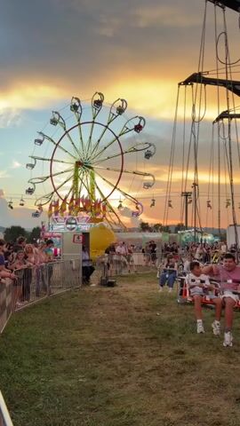 The video shows a crowded carnival scene during sunset. A large Ferris wheel with brightly colored lights is visible in the background, with gondolas ascending and descending. In the foreground, a swing ride is in motion, with two young boys seated on one of the swings, their arms raised. Several other people are seated on swings, and many more are gathered around watching the rides or walking through the carnival grounds. Fencing separates the main viewing area from the rides. The sky is filled with warm, vibrant colors from the sunset, casting a golden glow over the scene. The overall atmosphere appears festive and lively.