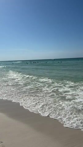 The video shows a beautiful beach with clear turquoise water and a bright blue sky. Small waves gently roll onto the sandy shore, creating a foamy white surf. In the distance, several people are visible swimming and enjoying the ocean. The overall atmosphere is peaceful and serene, capturing the essence of a perfect beach day.