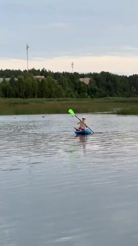 A person in a blue and green kayak paddles on a calm body of water. The person is wearing a light-colored shirt, shorts, and a hat. They are holding a paddle with bright green ends. In the background, there is a shoreline with tall grasses and trees. There are also some buildings and cell towers visible in the distance. The sky is overcast. The water is rippling slightly. There are a few birds flying in the distance.