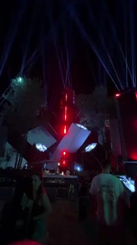 The video shows a crowded outdoor event at night.  A large, abstract stage structure is illuminated with red and blue lights, with beams of blue light projecting upwards into the dark sky.  Several trees surround the stage. In the foreground, people are visible standing and walking.  Some individuals are silhouetted against the bright stage lights.  A metal barrier separates the crowd from the stage area. The ground appears to be dirt or grass. The overall atmosphere suggests a music festival or concert.