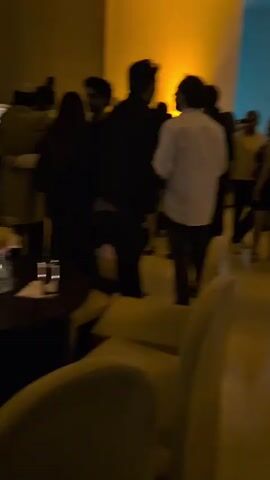 The video shows a gathering of people indoors. Most are standing and some are talking. The room appears to have a high ceiling, light-colored flooring, and glass walls. There are chairs and tables visible in the room. The lighting shifts from warm to cooler tones.