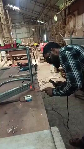 A man is welding a metal frame in a workshop. He is wearing a plaid shirt, welding mask, and gloves. Sparks are flying from the welding torch. In the background, there are various tools and materials, including a power drill, a level, and pieces of wood. The workshop appears to be a busy and functional space.