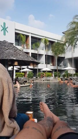 The video shows a person sitting by a pool, looking at a hotel building with palm trees. The person is wearing shorts and has tattoos on their legs. The pool is full of people swimming and relaxing. There are white umbrellas and plants around the pool. The sky is blue with some clouds.