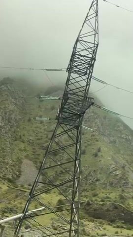 The video pans across a mountainous landscape partially obscured by fog. In the foreground, a large metal power line tower stands tall, with wires extending upwards and across the frame. Below the power line, a large pipe supported by concrete pillars runs horizontally across the scene. The mountains in the background are covered in green vegetation and rocky patches, with fog rolling down the slopes. The overall atmosphere is somewhat hazy and overcast, suggesting a cool, damp environment. There are no people or animals visible in the video. The movement of the camera is smooth and steady, indicating it is likely mounted on a moving vehicle.