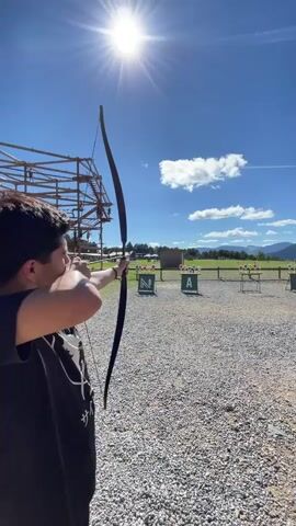 A person in a black t-shirt is shown from the side, aiming a bow and arrow at targets in the distance. The sun is shining brightly in a clear blue sky with a few white clouds. In the background, there is a wooden structure that appears to be part of an adventure park or obstacle course. Several targets, marked with letters and colorful circles, are set up on a gravel field. The person is in the process of drawing the bowstring, preparing to release an arrow.