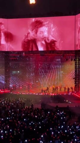 The video shows a live concert with a large crowd in attendance. On a brightly lit stage, performers are visible. Above the stage, a large screen displays close-up shots of the performers, with red lighting dominating the background. The crowd is holding up lights, seemingly from phones or light sticks, which appear as bright dots in the darkened audience area. Steel beams and scaffolding structure is visible around the stage setup.